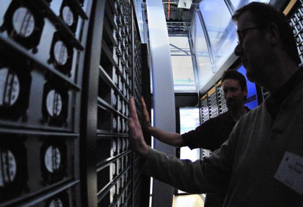 Interior of a data center hot aisle with rows of server racks and two people inspecting equipment, illustrating large-scale server infrastructure.