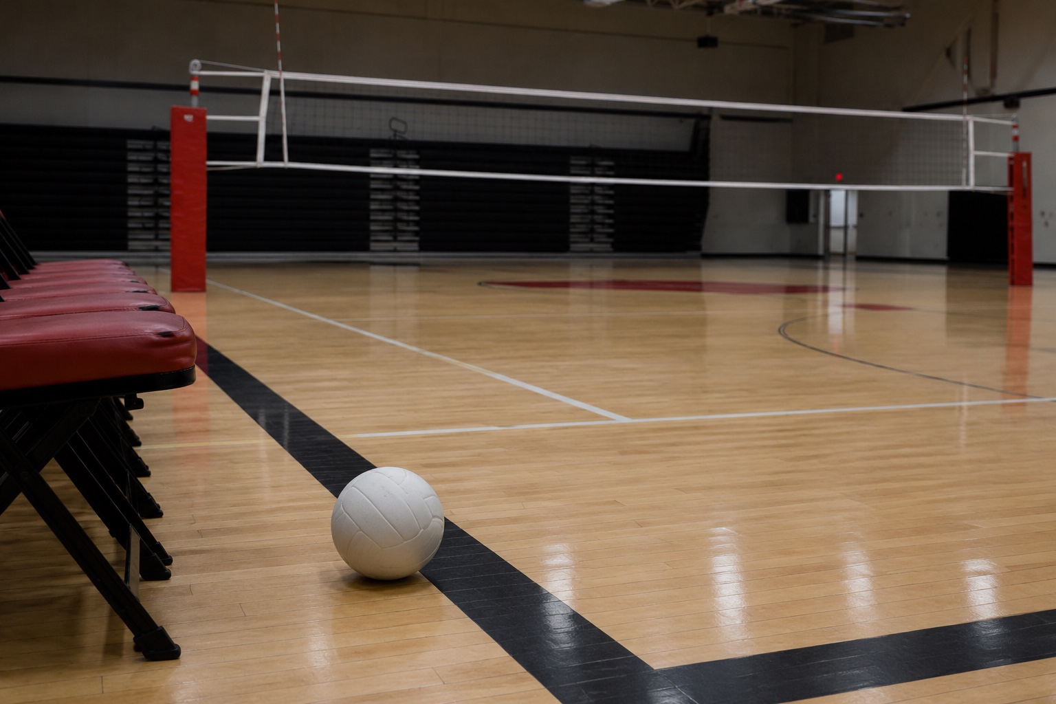 An empty high-school volleyball gym with a volleyball resting beside the team bench after a match.