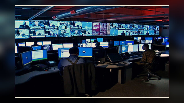 Interior view of a Defense Intelligence Agency 24/7 watch center with analysts at computer consoles and large displays on the walls.