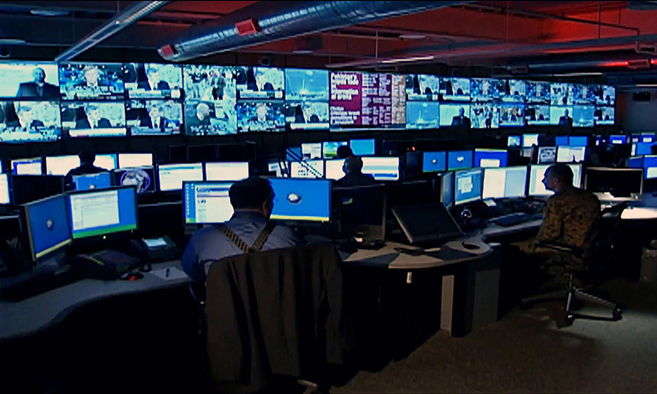 Interior view of a Defense Intelligence Agency 24/7 watch center with analysts at computer consoles and large displays on the walls.